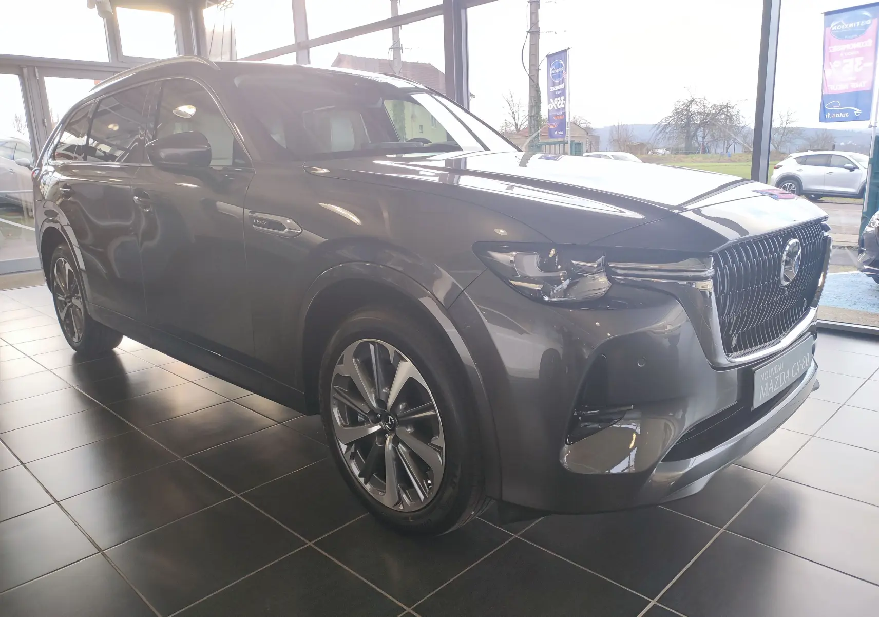 Vue 3/4 avant droit d'un Mazda CX-80 gris foncé hybride rechargeable exposé en showroom avec calandre imposante.