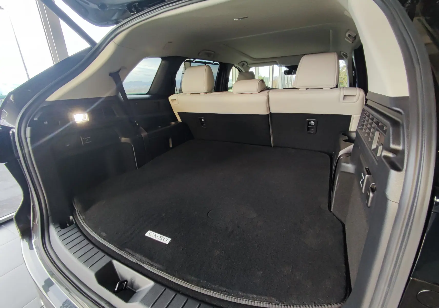 Vue arrière ouverte du coffre spacieux du Mazda CX-80 gris foncé avec banquette arrière beige visible.