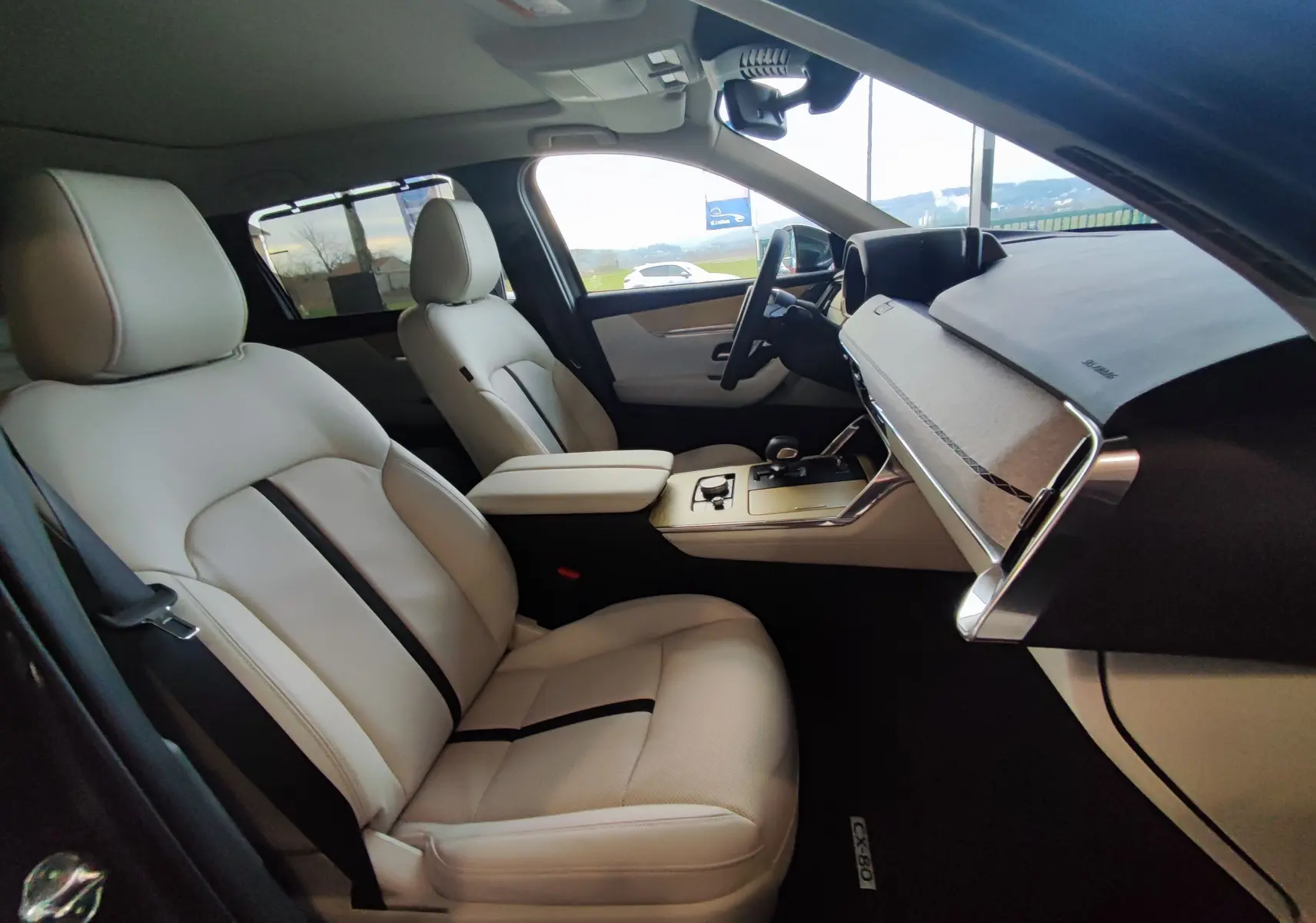Intérieur clair du Mazda CX-80 2024 vu côté conducteur, sièges cuir beige avec bande noire et tableau de bord moderne.