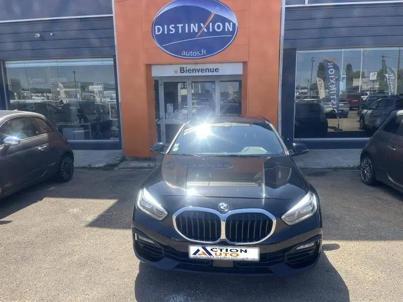 BMW Série 1 noire vue de face, calandre chromée et phares LED allumés devant un garage Distinxion
