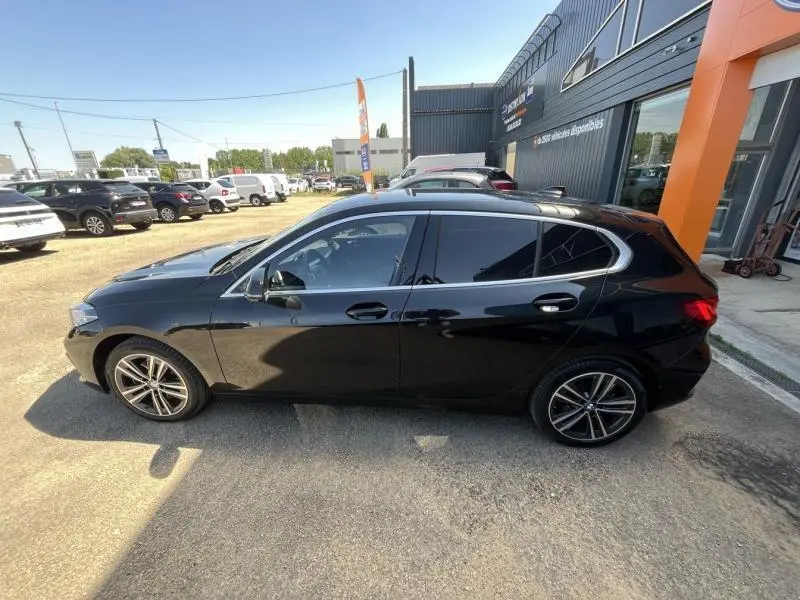 Profil côté gauche d'une BMW Série 1 116dA Lounge noire garée devant un bâtiment commercial.