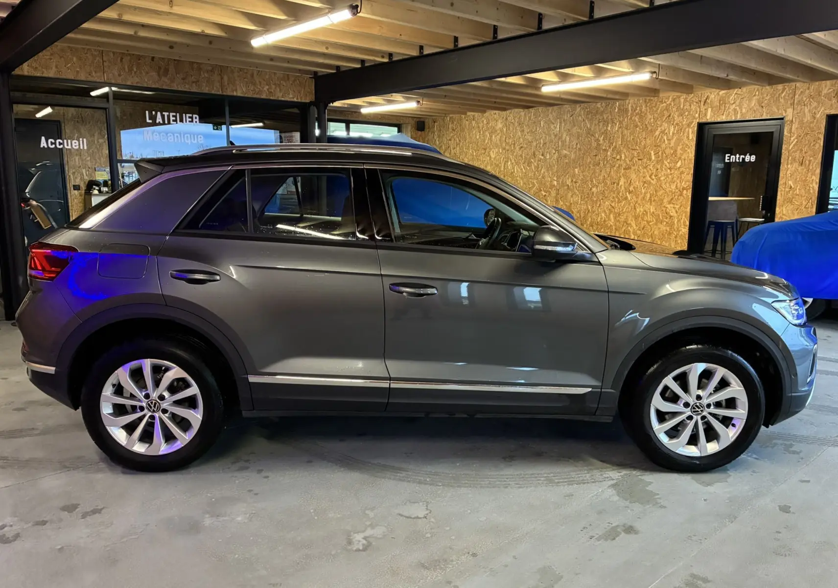 Vue de profil côté gauche d’un Volkswagen T-Roc gris 2023 garé en intérieur, avec jantes alliage et toit ouvrant.