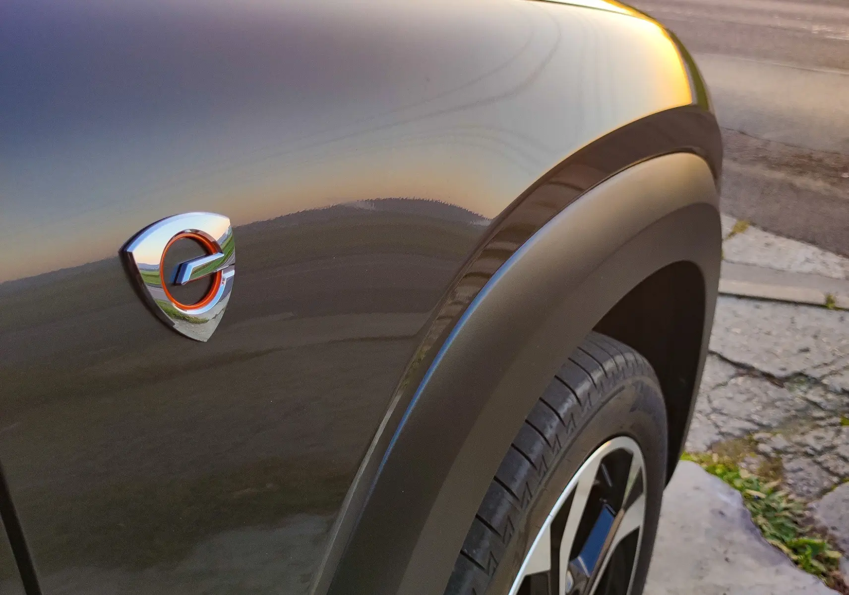 Gros plan sur l'aile avant droite gris foncé du Mazda MX-30 2023 avec logo hybride au coucher du soleil.