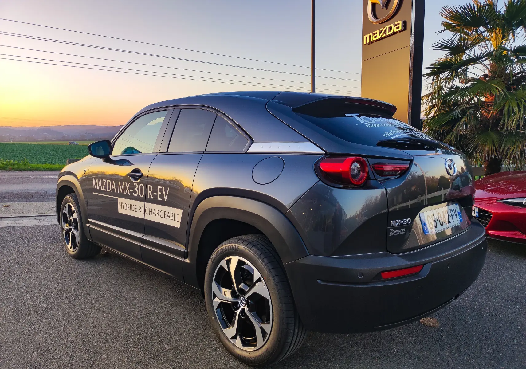Vue 3/4 arrière droite d'un Mazda MX-30 gris foncé hybride rechargeable avec logo Mazda et marquages visibles sur la porte.