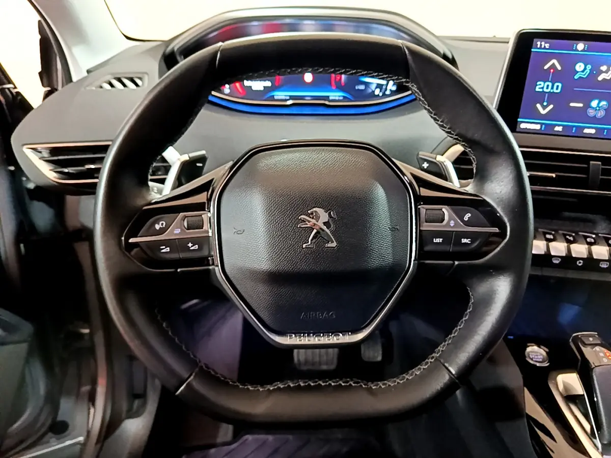 Volant cuir noir avec logo Peugeot, tableau de bord digital et écran tactile de climatisation dans un 5008 gris clair, vue intérieure frontale.