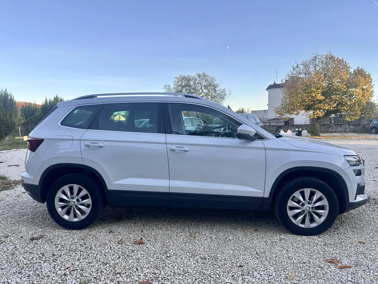 Vue de profil côté gauche d'un Skoda Karoq blanc 2024 sur un sol gravillonné en extérieur.