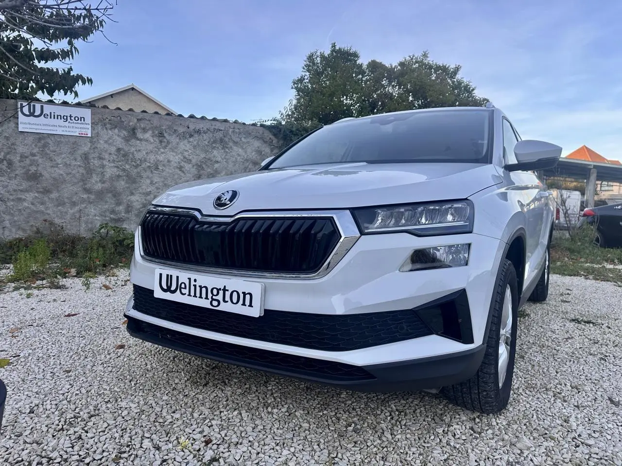 Vue 3/4 avant d'un Skoda Karoq blanc 2024 avec calandre noire et phares LED, sur un sol gravillonné.