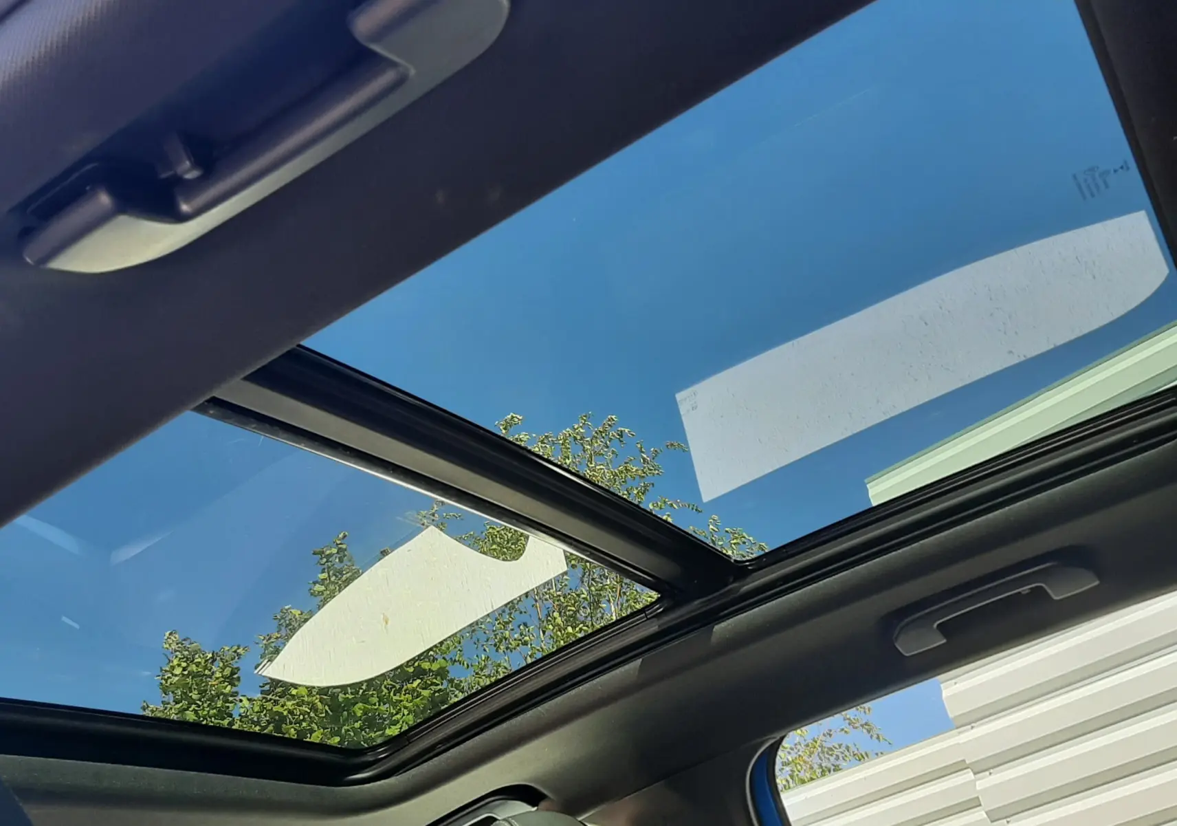 Toit panoramique ouvert vu de l'intérieur du Peugeot 3008 bleu Vertigo avec ciel bleu et arbres en arrière-plan.