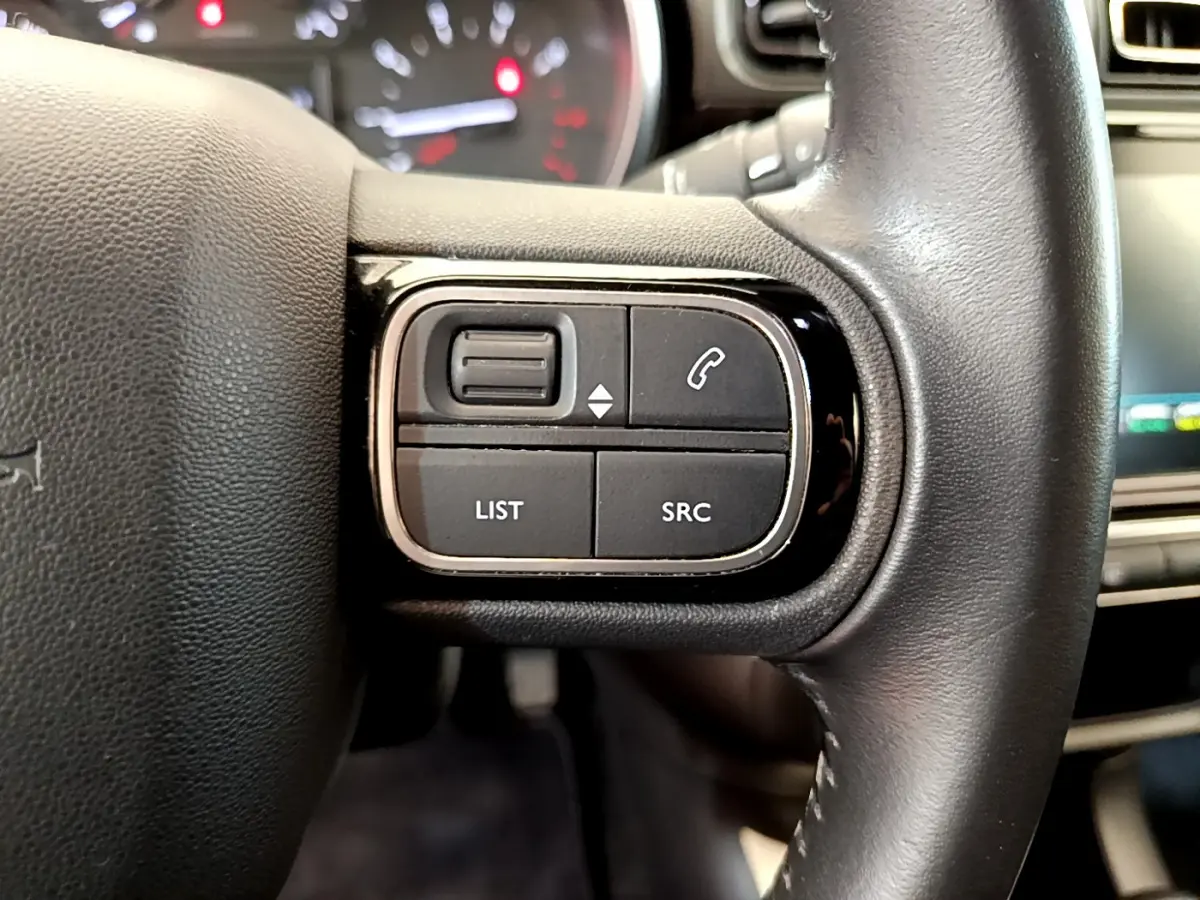Gros plan sur les commandes gauche du volant cuir noir d'une Citroën C3 gris clair, avec tableau de bord flou en arrière-plan.