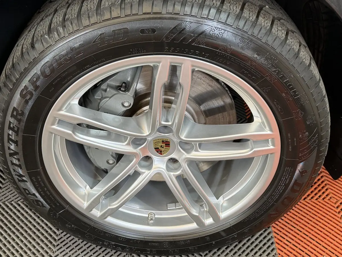 Gros plan sur la jante alu argentée avec logo Porsche centrale et pneu hiver Dunlop sur Porsche Macan gris foncé.
