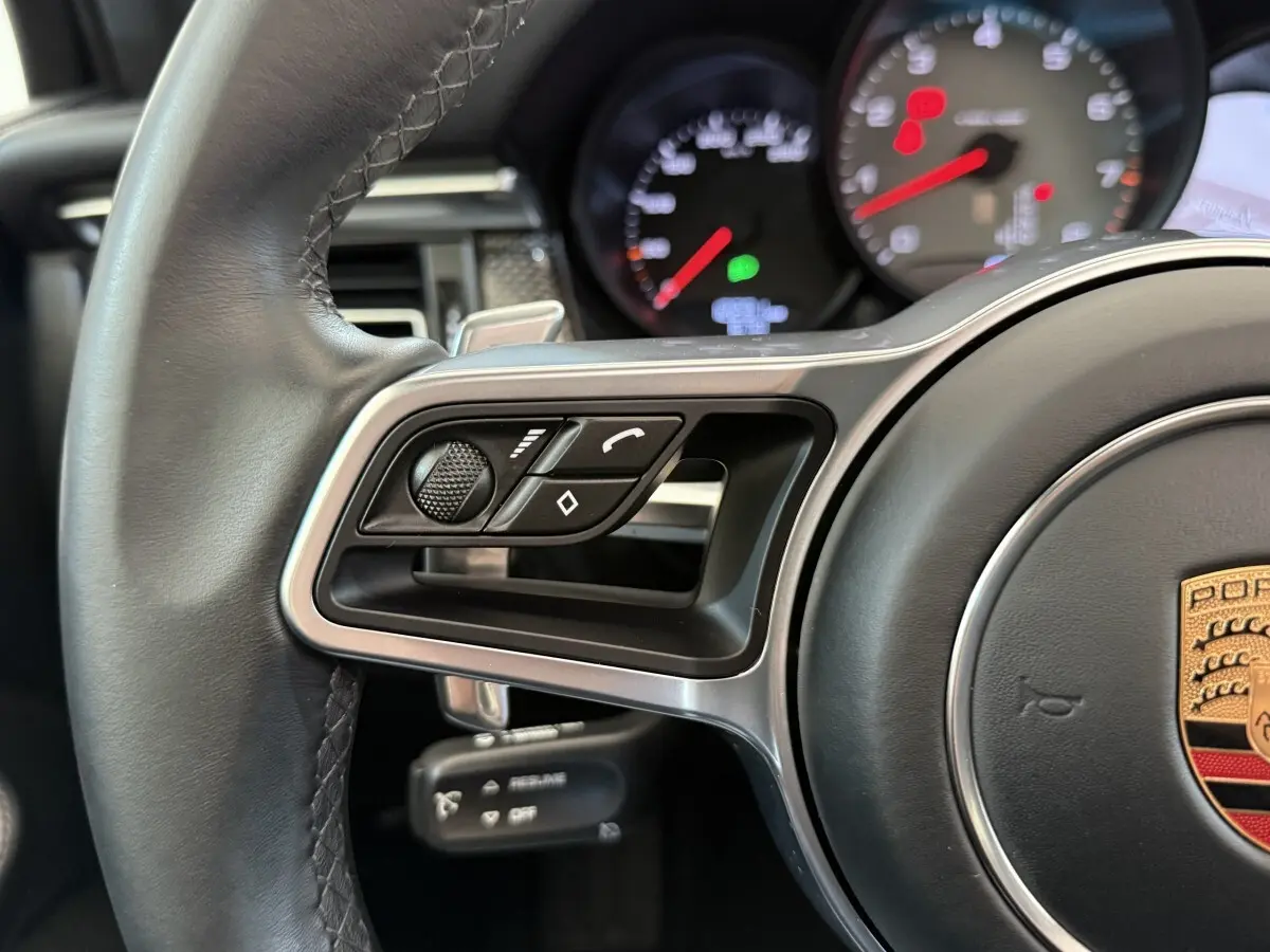 Gros plan sur le volant en cuir et alu du Porsche Macan 3.0 S, avec commandes multifonctions et tableau de bord partiellement visible.