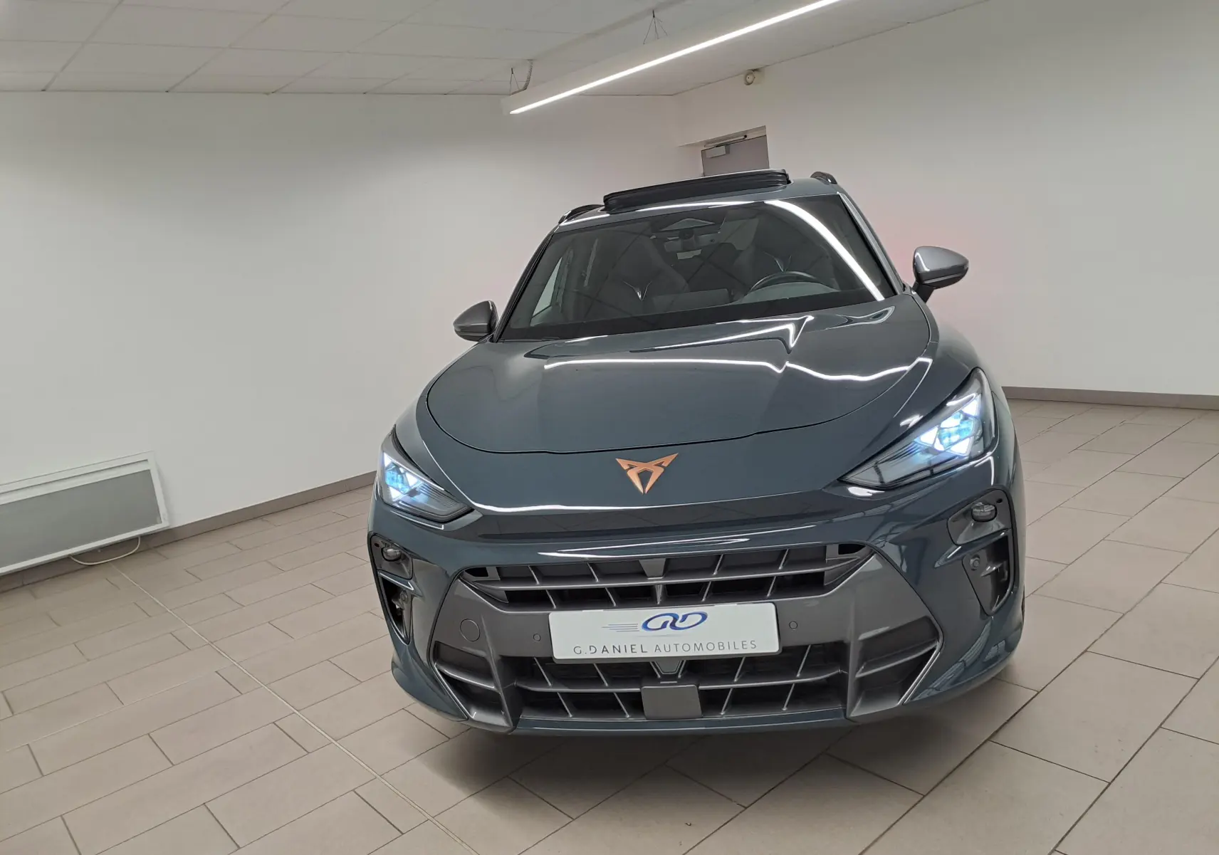 Vue avant d'une CUPRA TERRAMAR 2025 bleu fjord avec phares allumés et toit ouvrant panoramique visible en intérieur showroom.