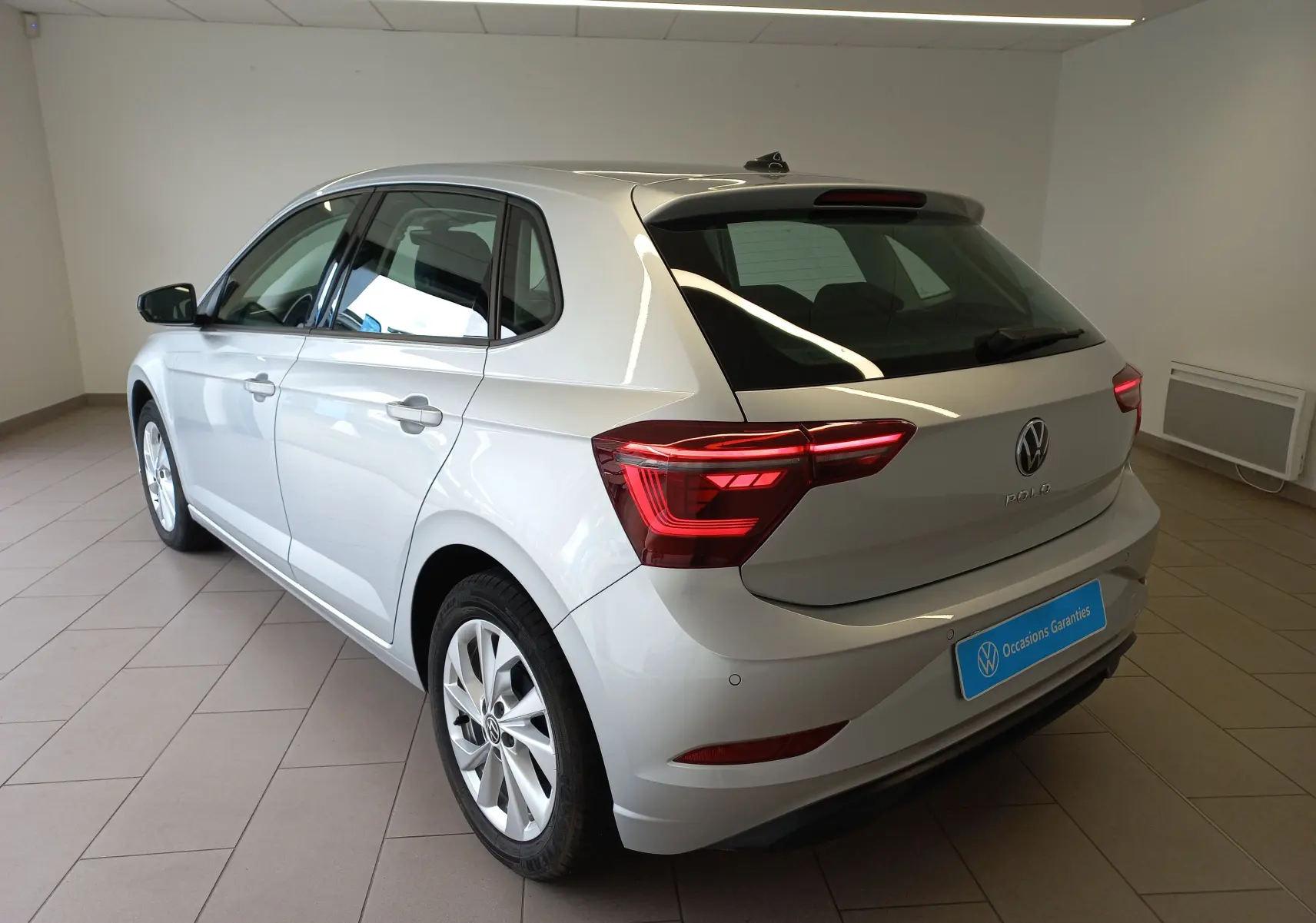 Vue 3/4 arrière droite d'une Volkswagen Polo 2026 argent reflet en intérieur, avec feux arrière allumés.