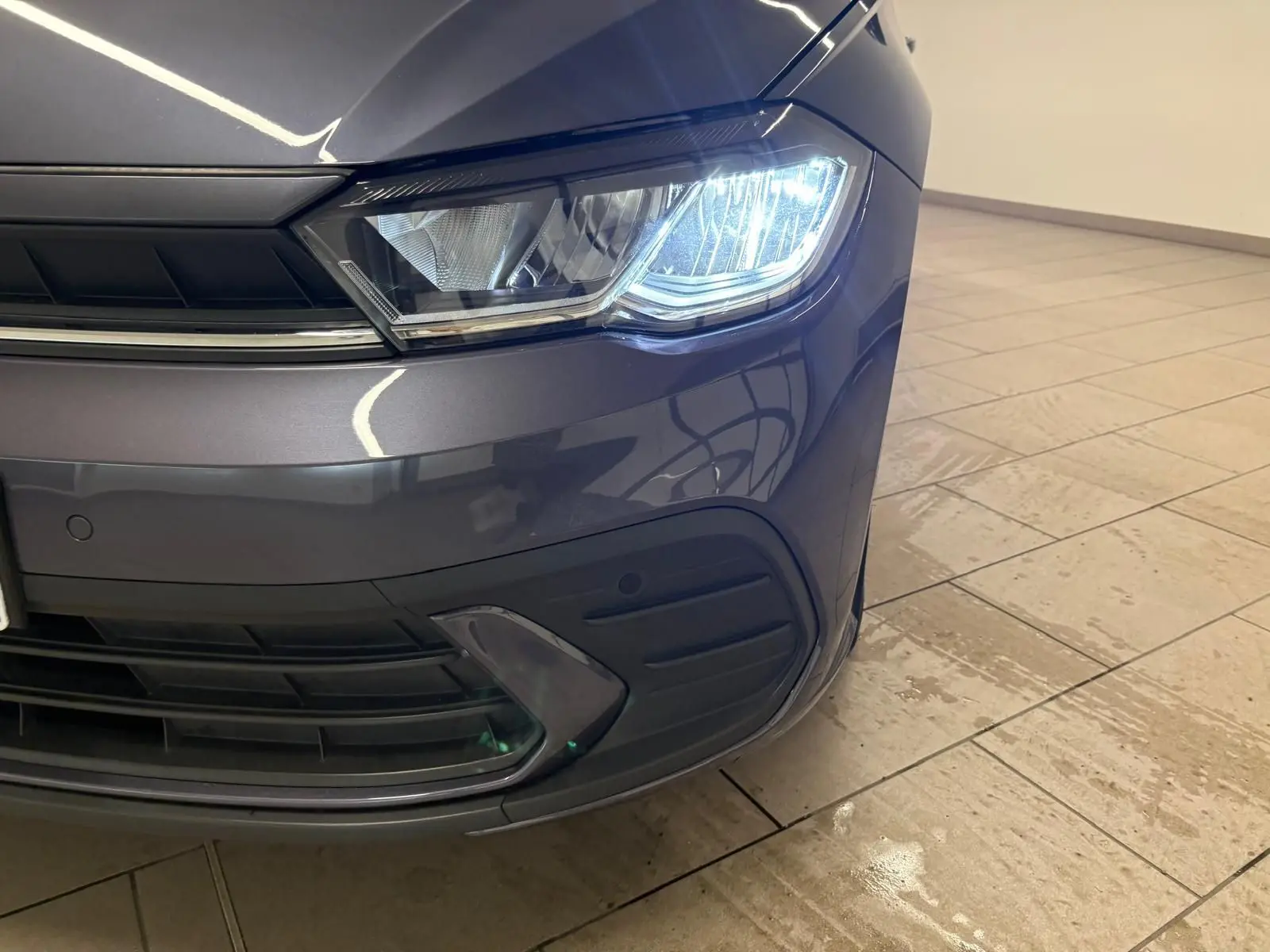 Gros plan sur le phare avant droit allumé d'une Volkswagen Polo gris cendre 2024 dans un intérieur lumineux.