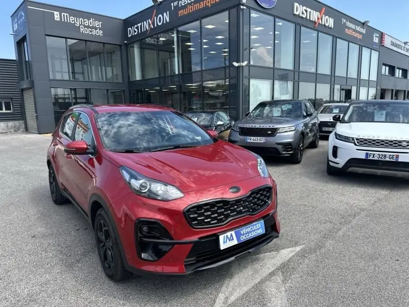 KIA SPORTAGE rouge métal vu en 3/4 avant droit, avec calandre noire et jantes foncées, devant un showroom multimarques.