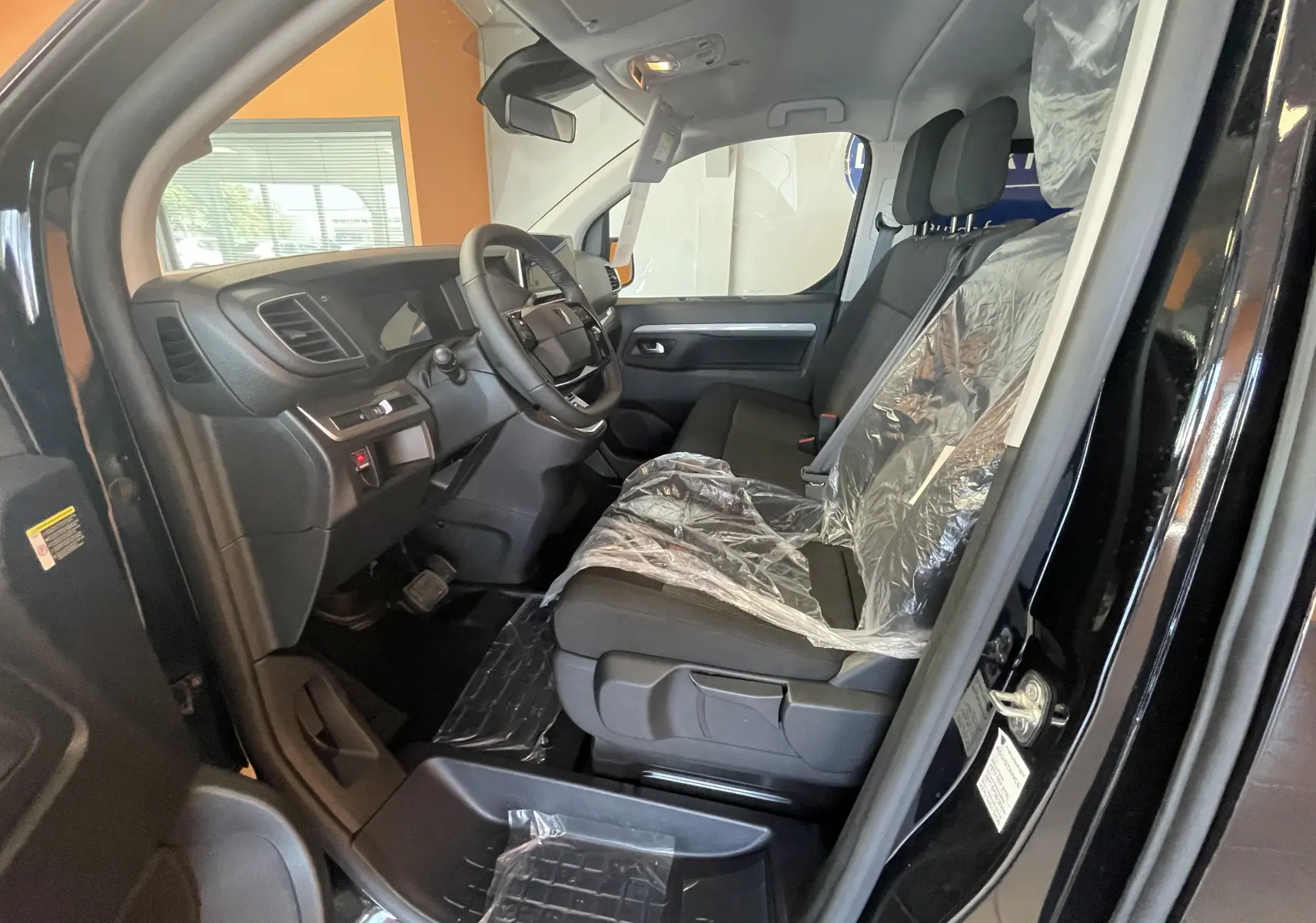 Vue intérieure côté conducteur du Peugeot Traveller noir 2026, montrant le tableau de bord et les sièges avant protégés par des housses plastiques.