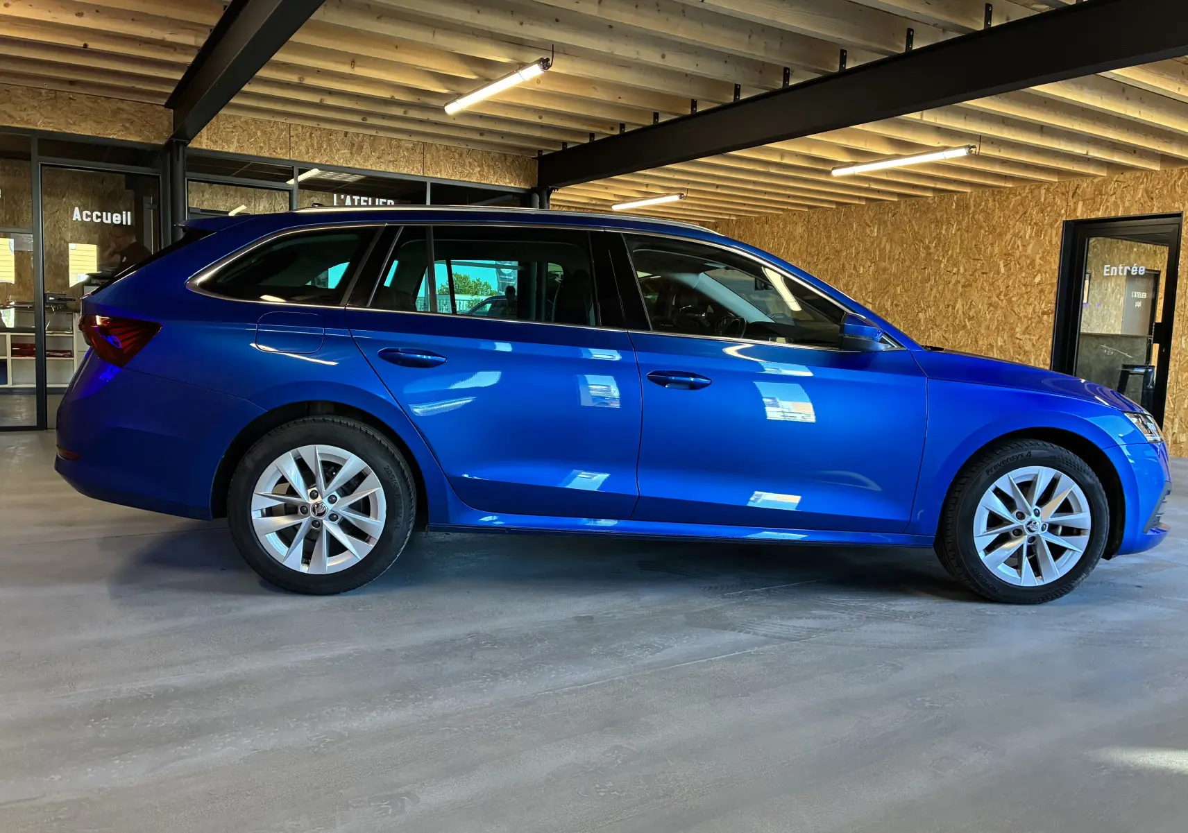 Vue latérale droite d'une Skoda Octavia Combi 2024 bleu brillant, stationnée en intérieur avec jantes alliage visibles.
