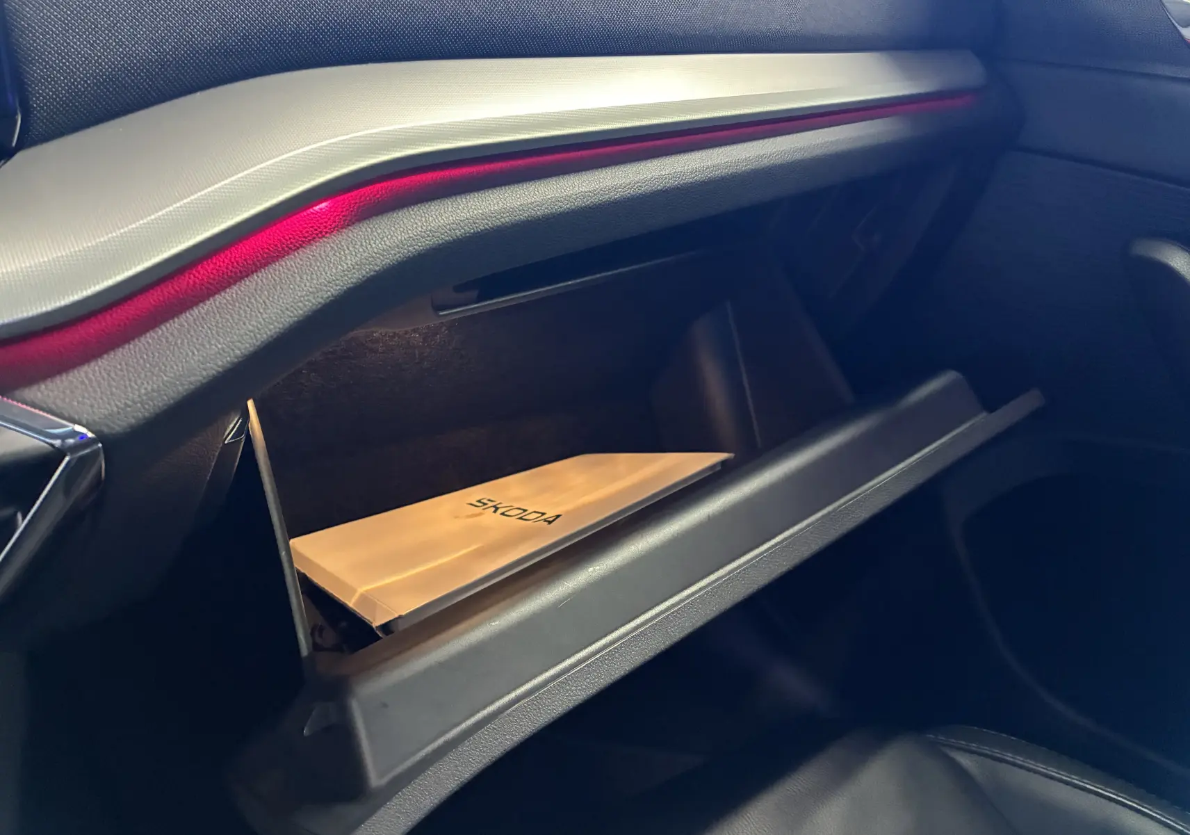 Gros plan sur la boîte à gants ouverte de la Skoda Octavia Combi 2024, avec un manuel Skoda visible et éclairage d’ambiance rouge.
