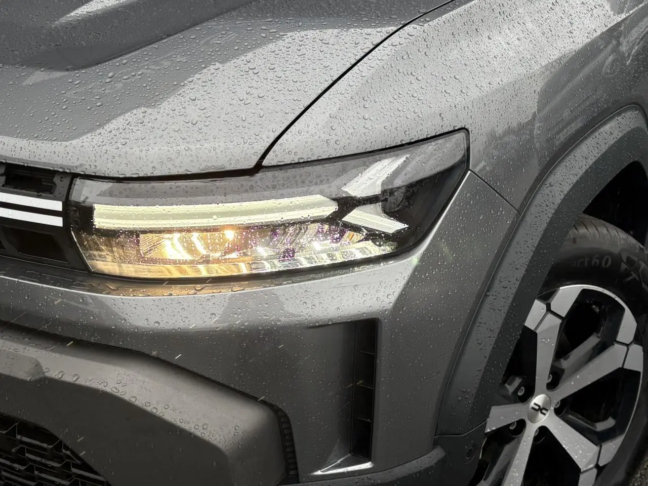 Gros plan sur l'avant droit gris schiste du Dacia Duster 2026 avec phare allumé et jante alliage diamantée.