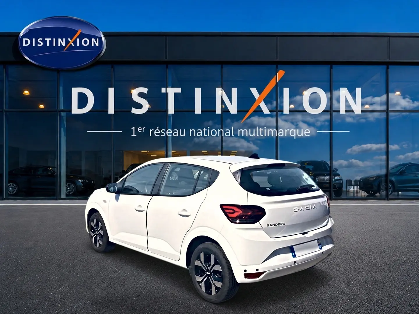 Vue 3/4 arrière droite d'une Dacia Sandero blanche 2025 avec jantes alu et feux arrière LED, devant un showroom Distinxion.