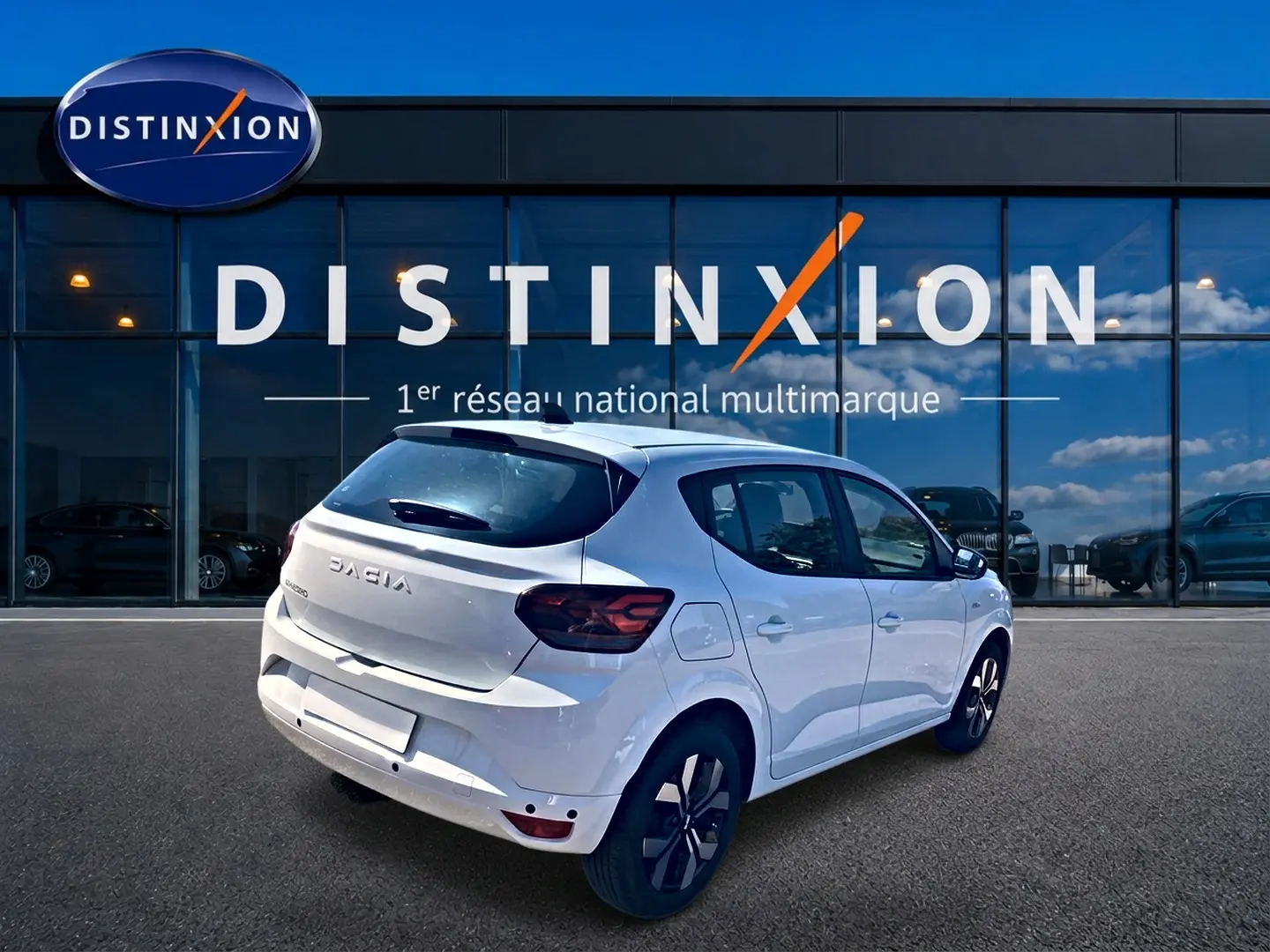 Vue 3/4 arrière droite d'une Dacia Sandero blanche 2025 avec jantes alu et becquet arrière, devant un showroom Distinxion.