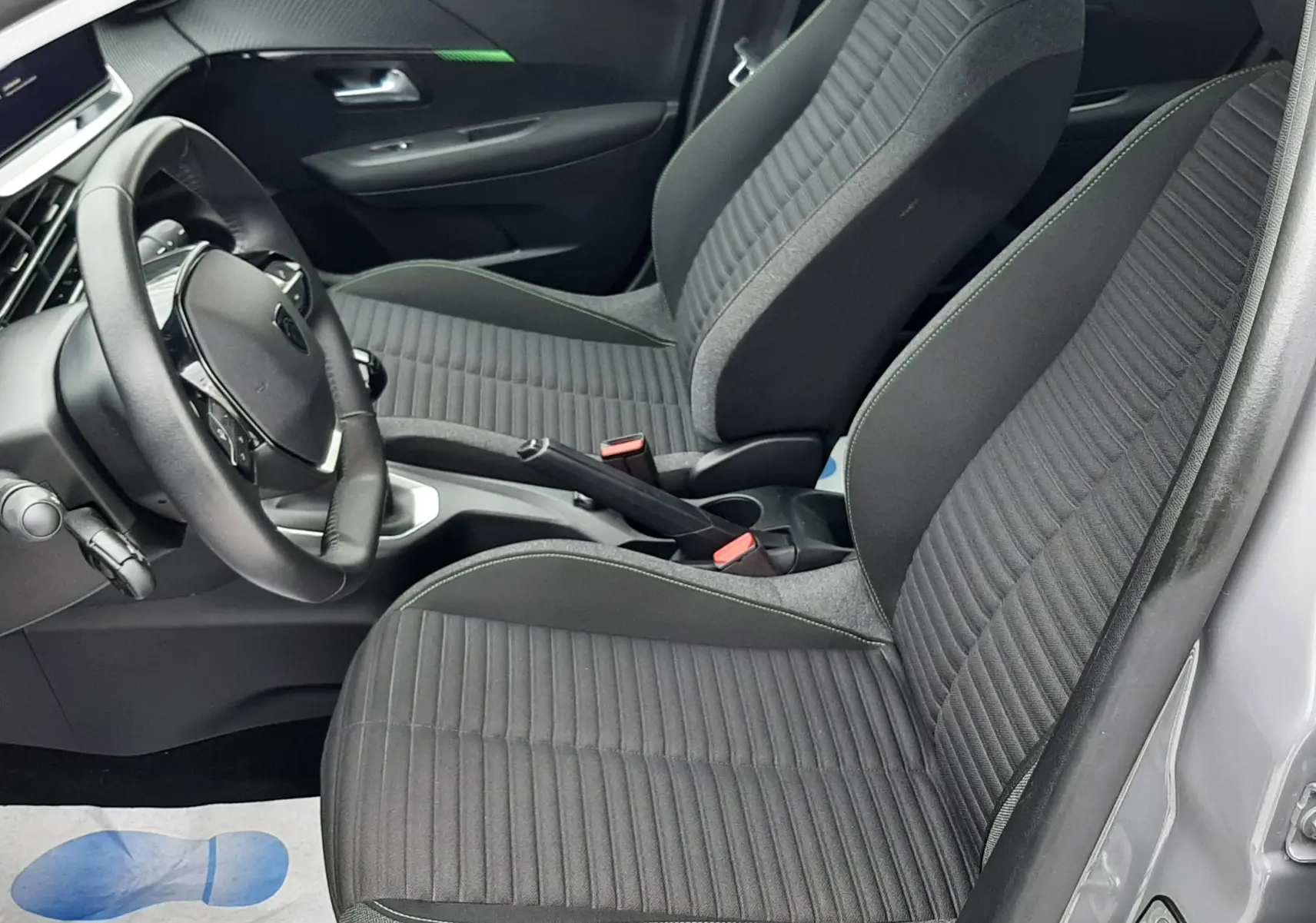 Vue intérieure côté conducteur de la Peugeot 208 2024 gris selenium, sièges tissu gris avec coutures vertes et volant multifonction.