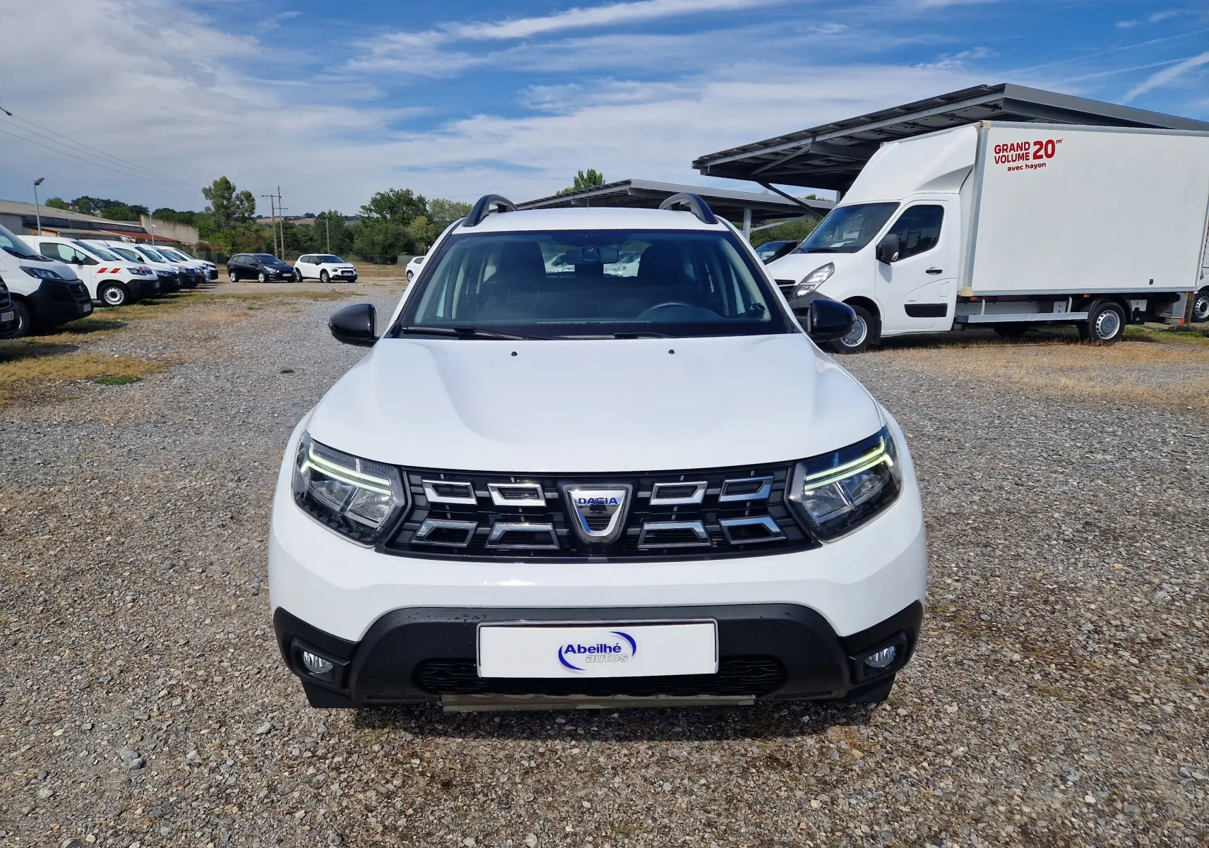 Vue frontale d'un Dacia Duster 4x4 blanc 2022 avec calandre noire et phares LED allumés sur un parking extérieur.