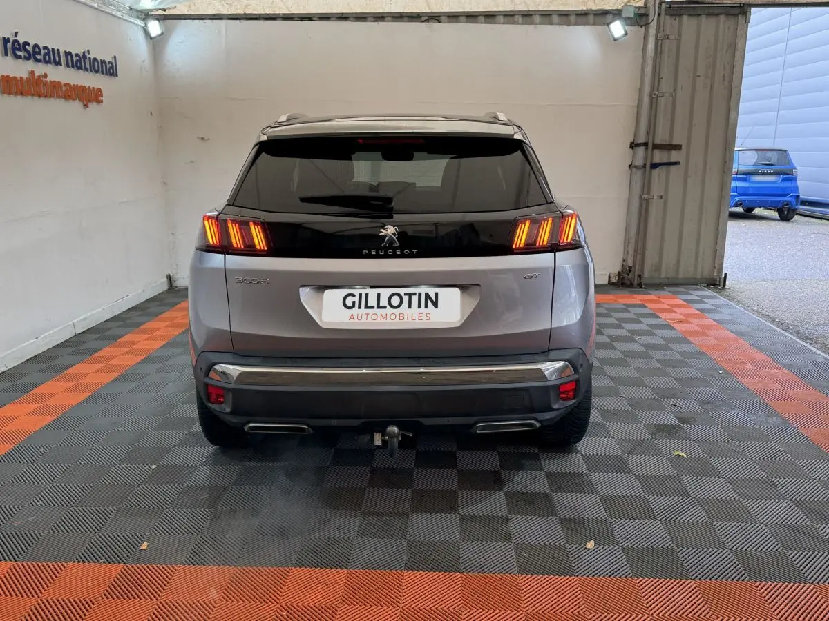 Vue arrière d'un Peugeot 3008 GT gris clair avec feux LED allumés et attelage visible en intérieur showroom.
