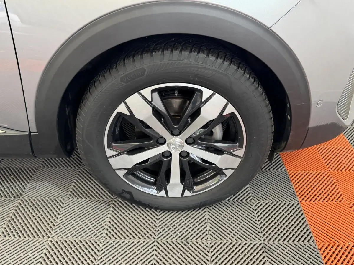 Gros plan sur la roue avant gauche du Peugeot 3008 gris clair avec jante alliage bicolore et logo Peugeot au centre.