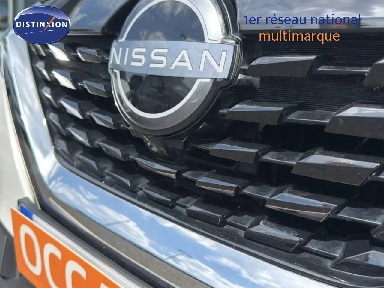 Gros plan sur la calandre noire et le logo Nissan d’un Qashqai blanc nacré 2023, avec reflet du ciel visible.