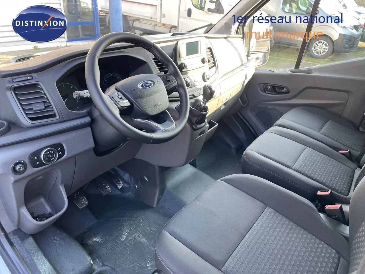Intérieur du Ford Transit Fourgon blanc 2023 vu côté conducteur, montrant tableau de bord et sièges noirs.