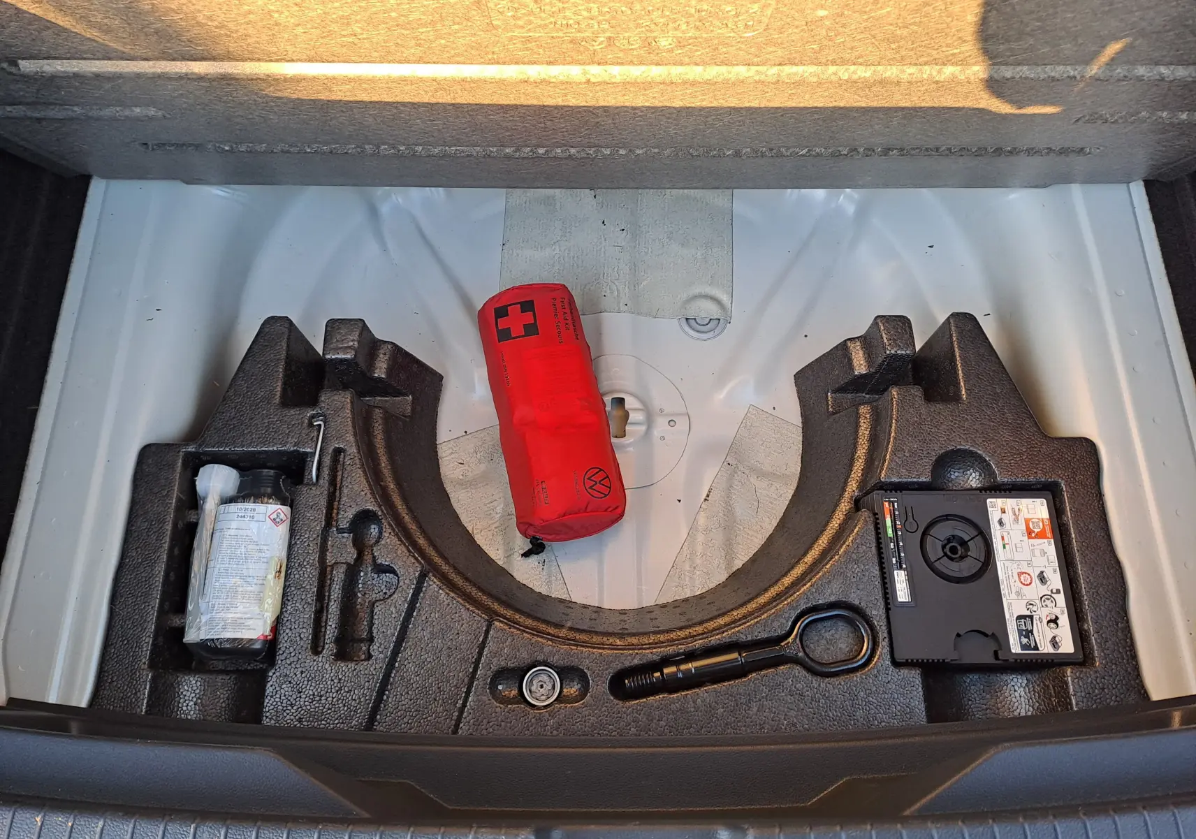 Coffre ouvert du Volkswagen T-Roc gris Ascot, montrant le rangement sous plancher avec kit de réparation et trousse de secours rouge.