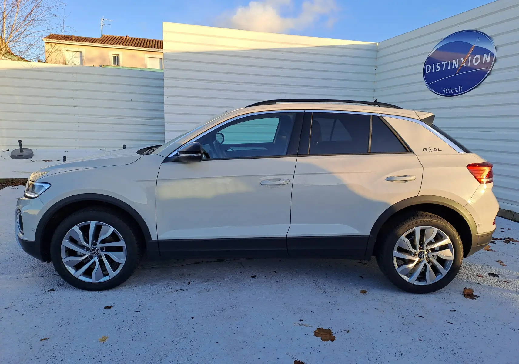 Vue latérale droite d'un Volkswagen T-Roc gris clair avec jantes alliage et toit noir, stationné devant un mur blanc.