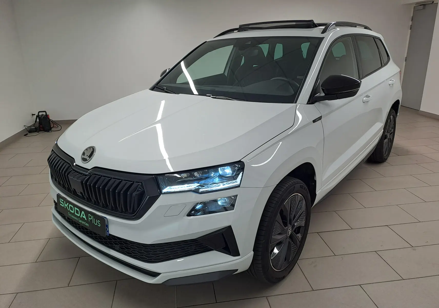 Skoda Karoq 1.5 TSI 150 DSG7 Sportline blanc lune en 3/4 avant droit, avec calandre noire et feux allumés.