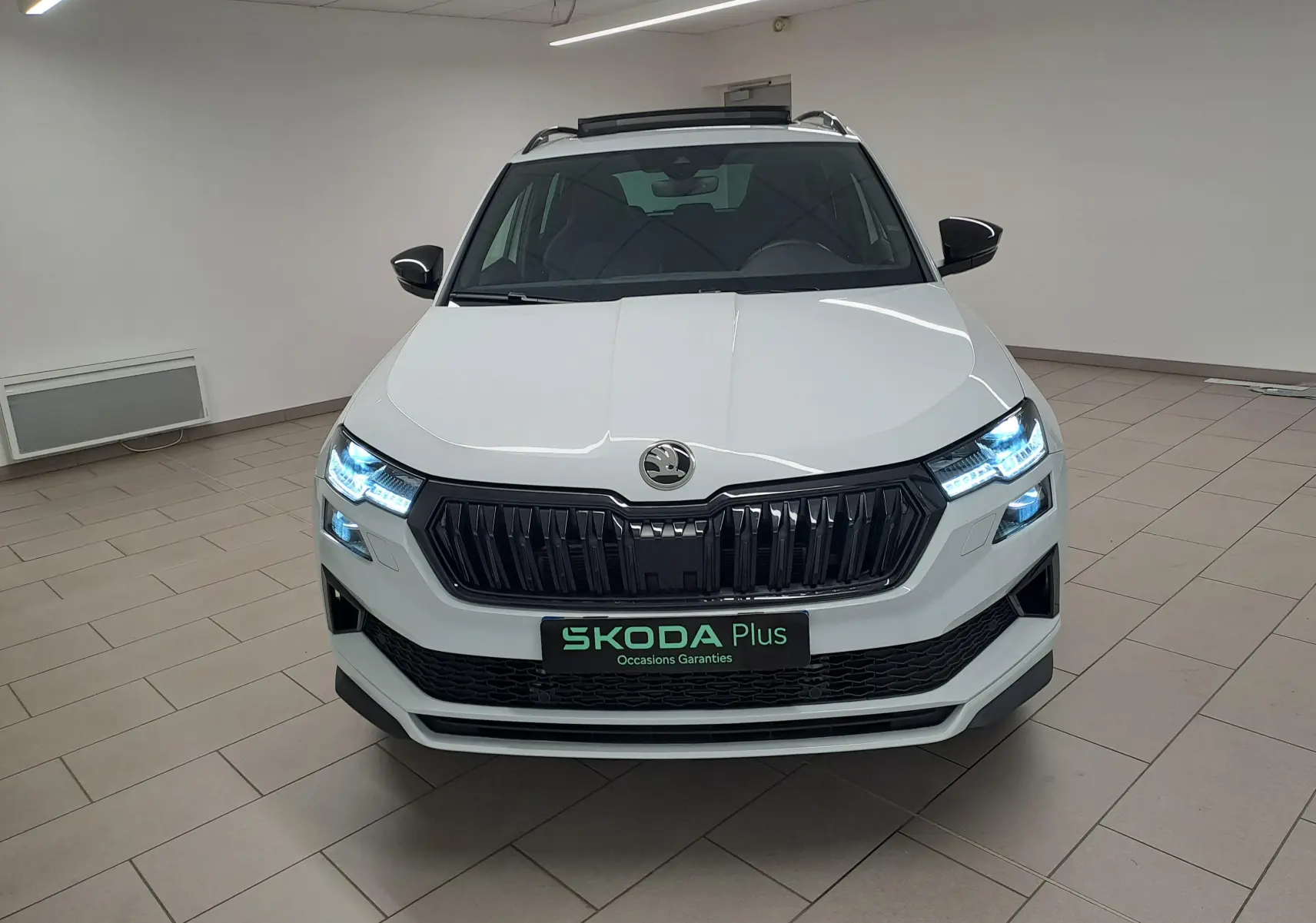 Vue frontale d’un Skoda Karoq Blanc Lune 2025 avec phares LED allumés et calandre noire Sportline.