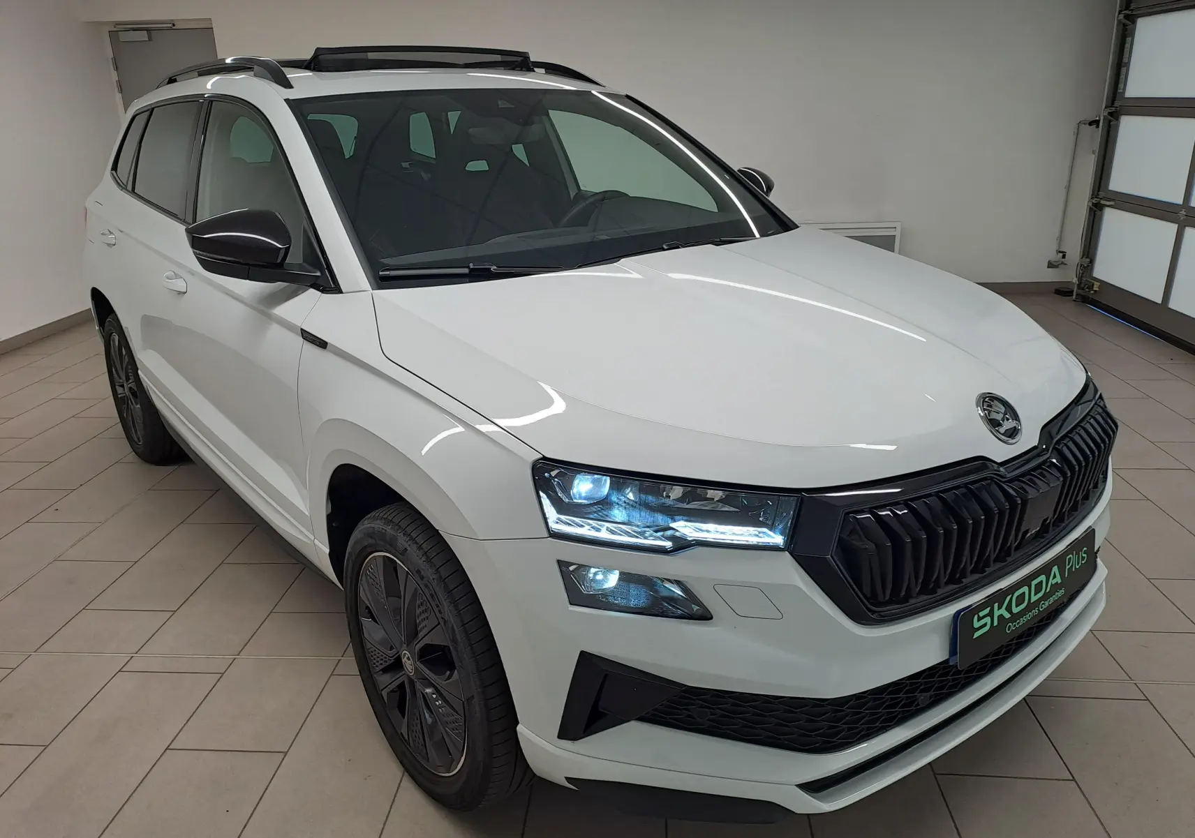 Vue 3/4 avant droite du Skoda Karoq Blanc Lune 2025 avec calandre noire et phares LED allumés en intérieur.