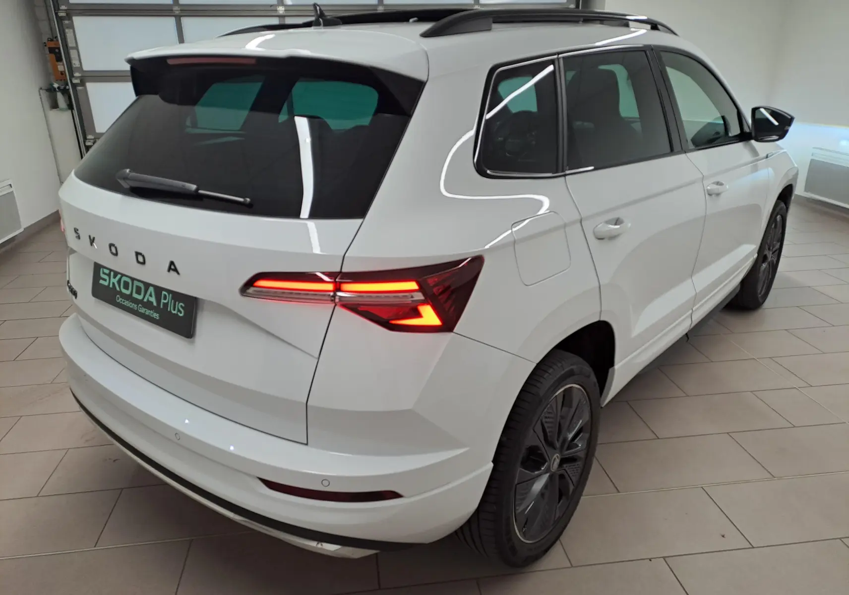 Vue 3/4 arrière droite d'un Skoda Karoq blanc lune 2025 avec feux arrière allumés et jantes noires sportline.