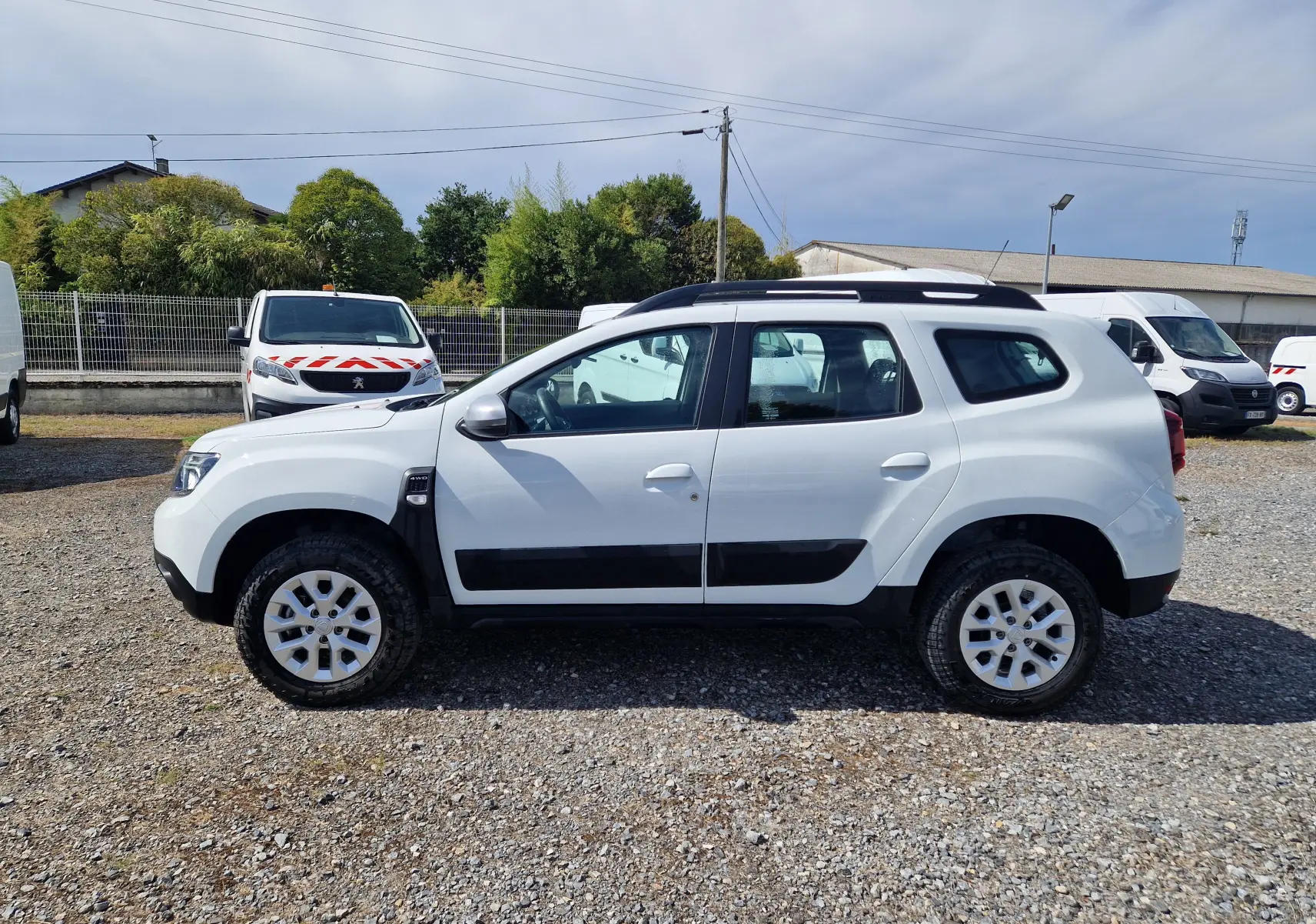 Profil côté gauche d'un Dacia Duster blanc 4x4 2023 avec protections noires et jantes alliage 16 pouces sur sol gravillonné.