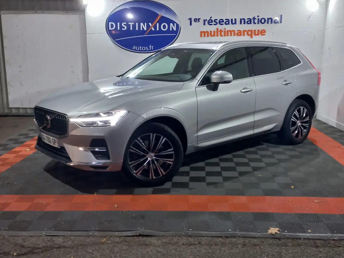 Volvo XC60 gris clair en 3/4 avant droit, stationné en intérieur sur sol damier noir et orange.