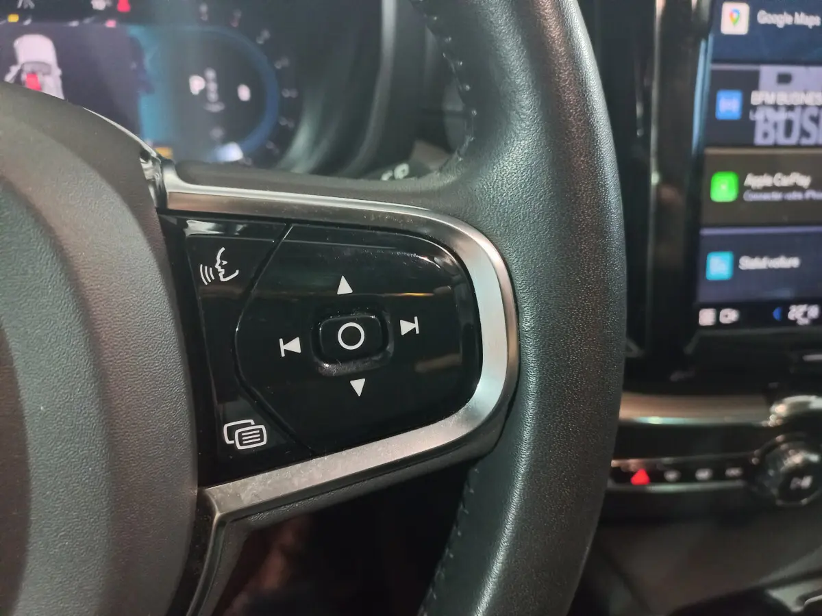 Gros plan sur les commandes vocales et multimédia du volant noir du Volvo XC60 B4 gris clair, avec écran tactile en arrière-plan.