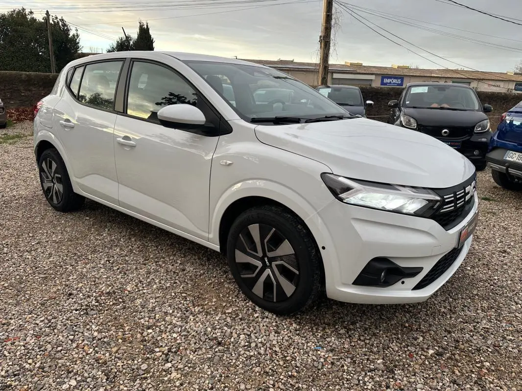 Dacia Sandero TCe 90 blanc vue 3/4 avant droit sur parking avec phares allumés et jantes stylisées.