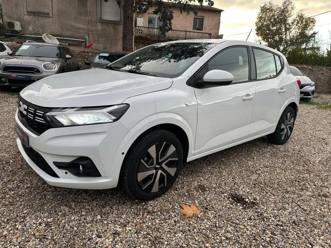 Vue 3/4 avant droite d'une Dacia Sandero TCe 90 Expression blanche avec jantes noires et phares allumés.