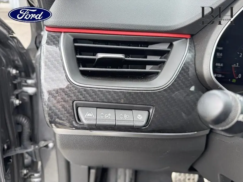 Détail de la console côté conducteur avec aérateur et boutons de réglages, finition carbone et liseré rouge visible.