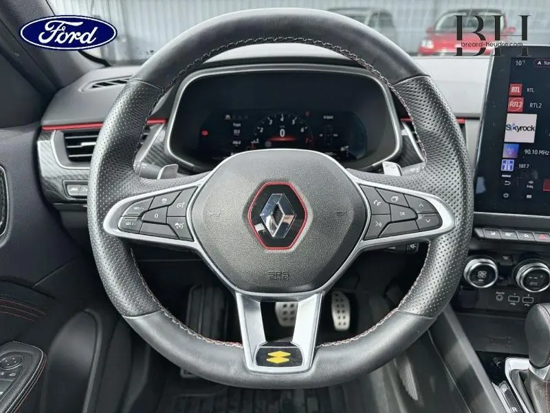 Vue rapprochée du volant cuir noir avec surpiqûres rouges du Renault Arkana gris métallisé, tableau de bord et écran tactile visibles.