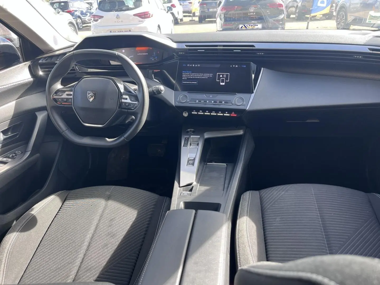 Vue intérieure avant de la Peugeot 308 2024, montrant le volant compact, l'écran tactile central et les sièges tissu gris.