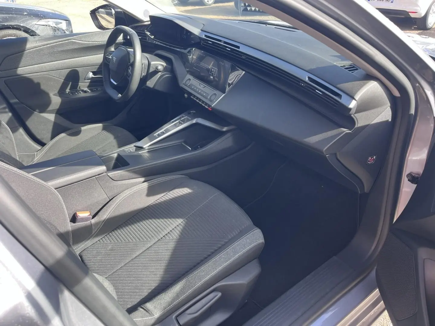 Vue intérieure côté conducteur de la Peugeot 308 2024 gris Artense, avec tableau de bord moderne et sièges tissu gris.