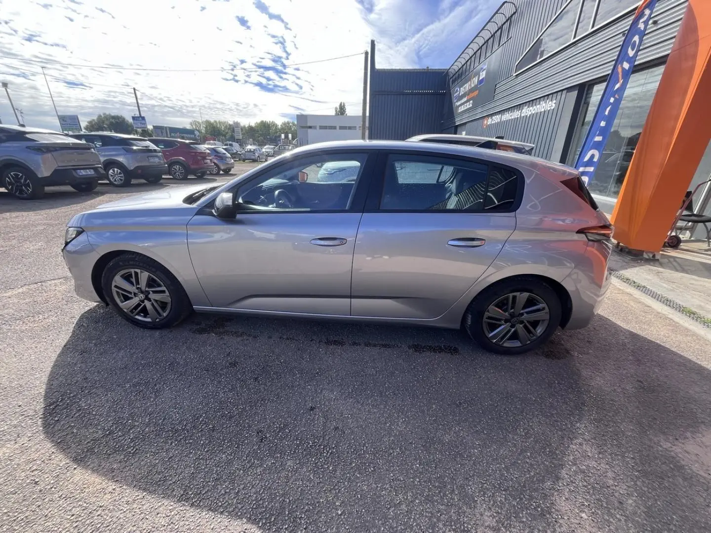 Peugeot 308 gris Artense vue de profil côté gauche, garée devant un bâtiment commercial.