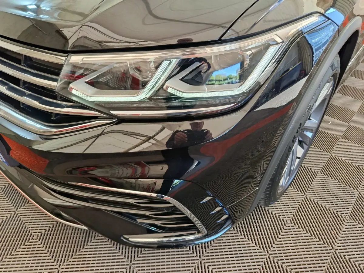 Gros plan sur le phare avant droit noir brillant du Volkswagen Tiguan 1.4 eHybrid 2023 avec détails chromés et jante visible.