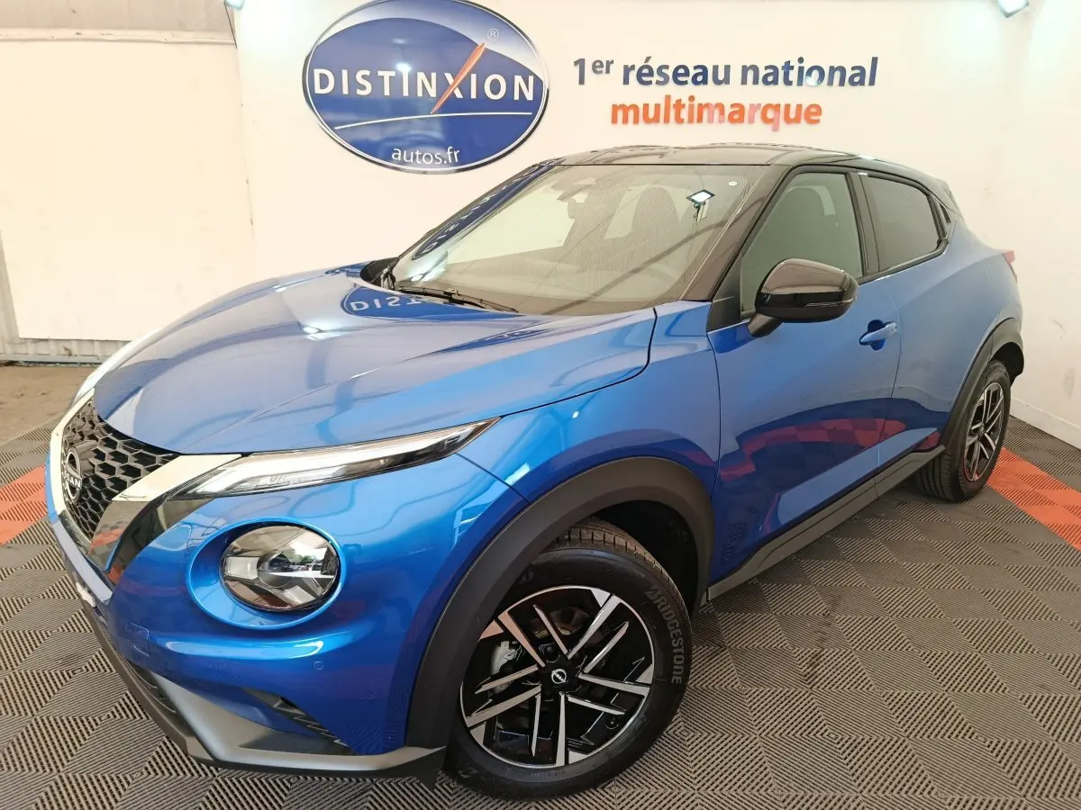 Nissan Juke bleu en 3/4 avant droit, avec toit noir et jantes alliage distinctives, en intérieur showroom.