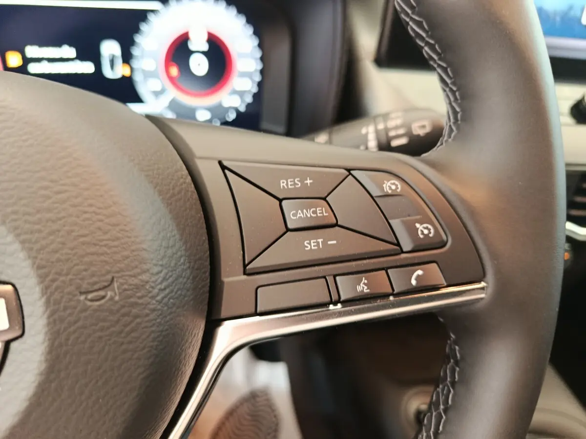 Gros plan sur les commandes multifonctions du volant cuir noir du Nissan Juke bleu 2025, avec tableau de bord numérique en arrière-plan.